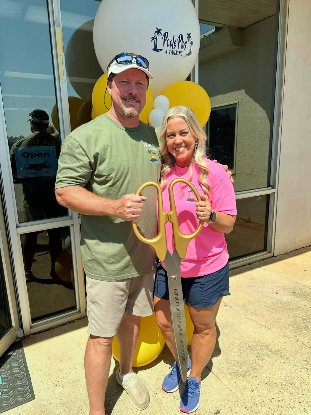 Two members of the Pools Plus and Tanning team holding oversized George County Chamber of Commerce scissors outside the shop